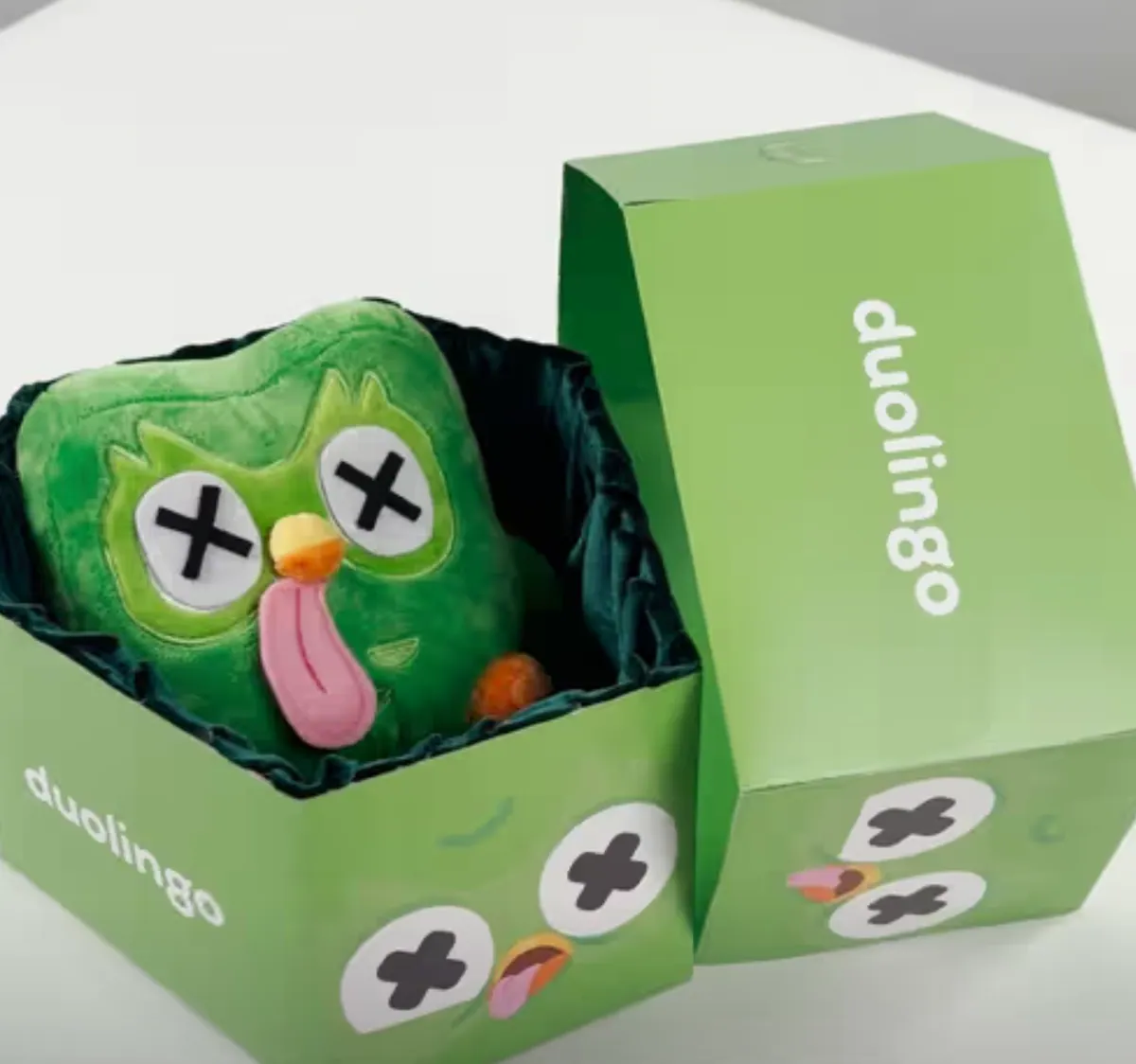 Peluche verde del gufo Duolingo con gli occhi a croce dentro una scatola regalo verde con il logo Duolingo