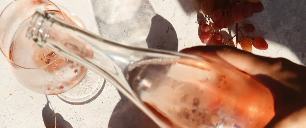Mano che versa vino rosato da una bottiglia in un calice, accanto a un grappolo d'uva