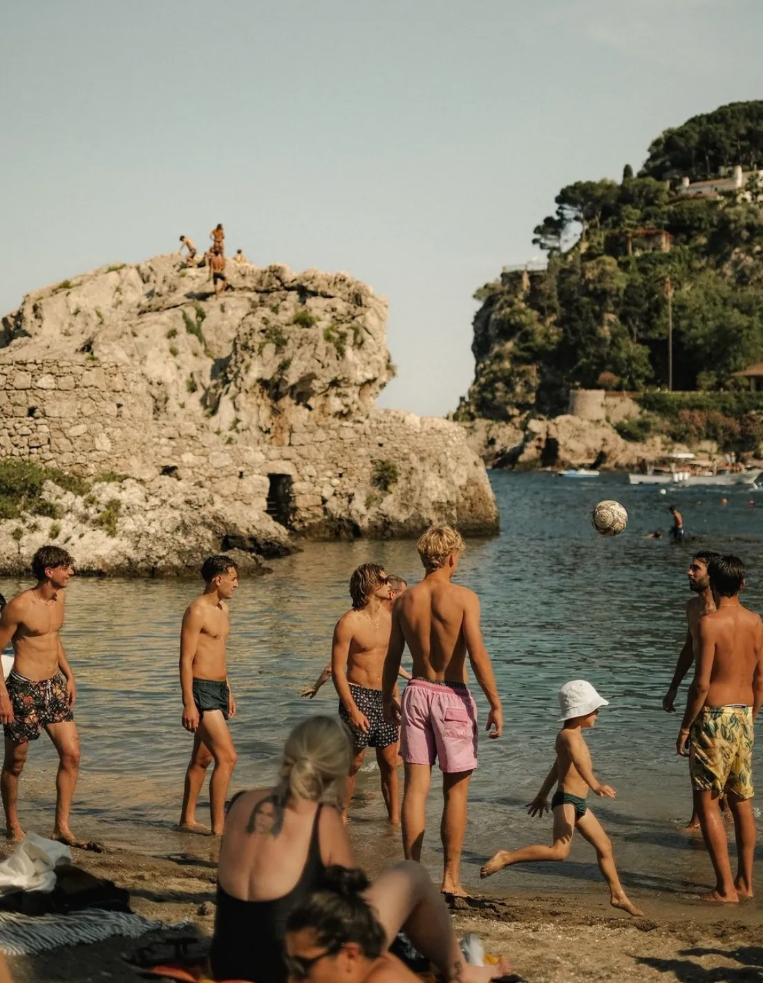 Young people playing football at the water's edge in front of a rocky islet on a Mediterranean beach