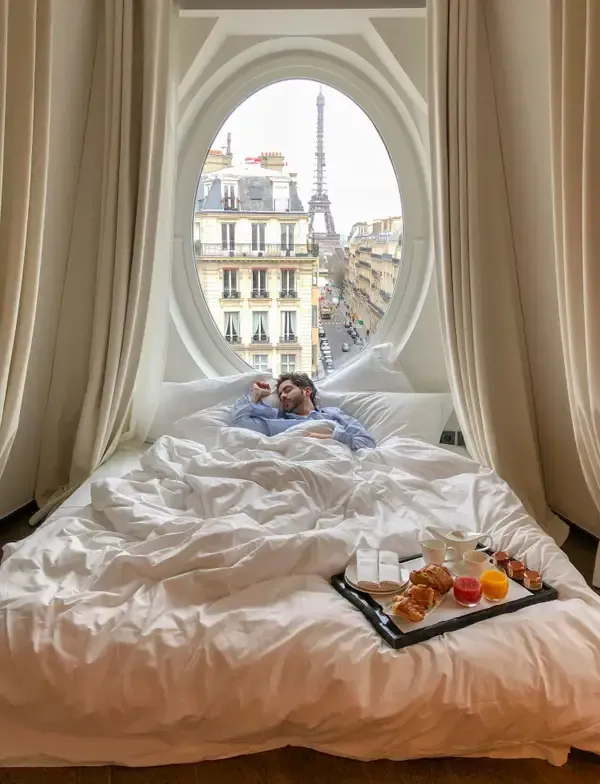 Uomo che dorme in un letto d'hotel con un vassoio della colazione, davanti a una finestra ovale con vista sulla Torre Eiffel