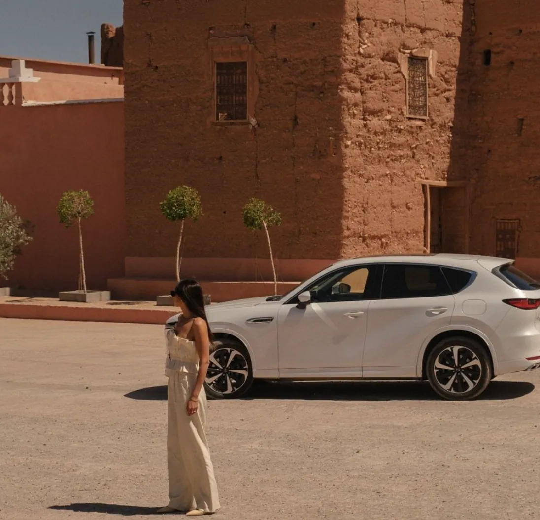 Donna in abito chiaro in piedi davanti a un SUV bianco, sullo sfondo di una kasbah in terra cruda