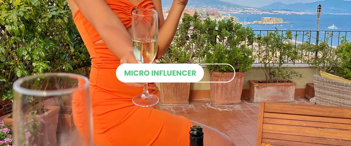 Una micro influencer in abito arancione con un calice di spumante su una terrazza affacciata sul golfo di Napoli, con la scritta Micro influencer