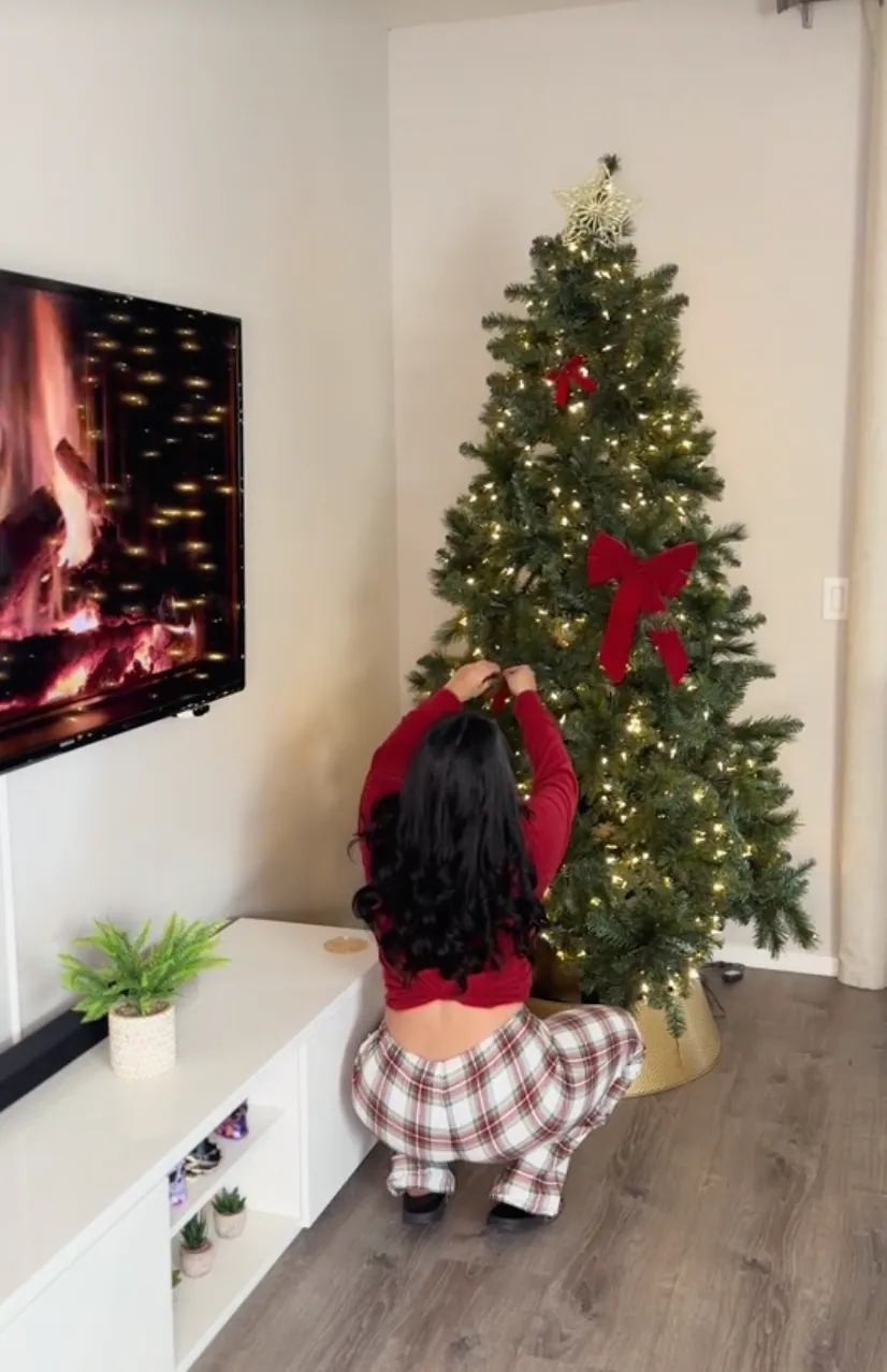 Ragazza in pigiama a quadri che decora un albero di Natale illuminato accanto a una TV con il camino acceso