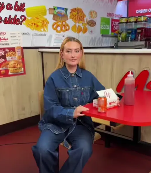 Una creator in giacca e jeans di denim seduta al tavolo rosso di un fast food, con una lattina e una confezione da asporto