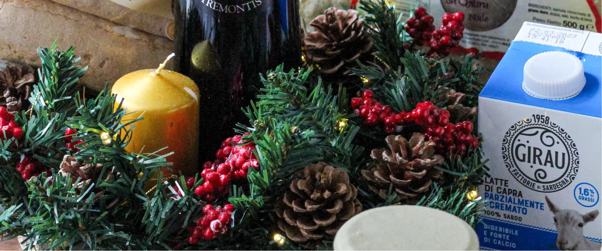 Composizione natalizia con rametti di abete, pigne, bacche rosse e una candela dorata accanto a una bottiglia di vino e a prodotti alimentari sardi