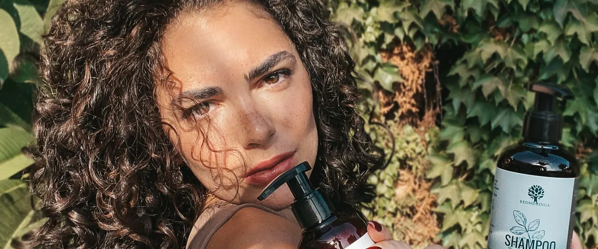 Curly-haired creator posing with a bottle of shampoo in front of an ivy-covered wall