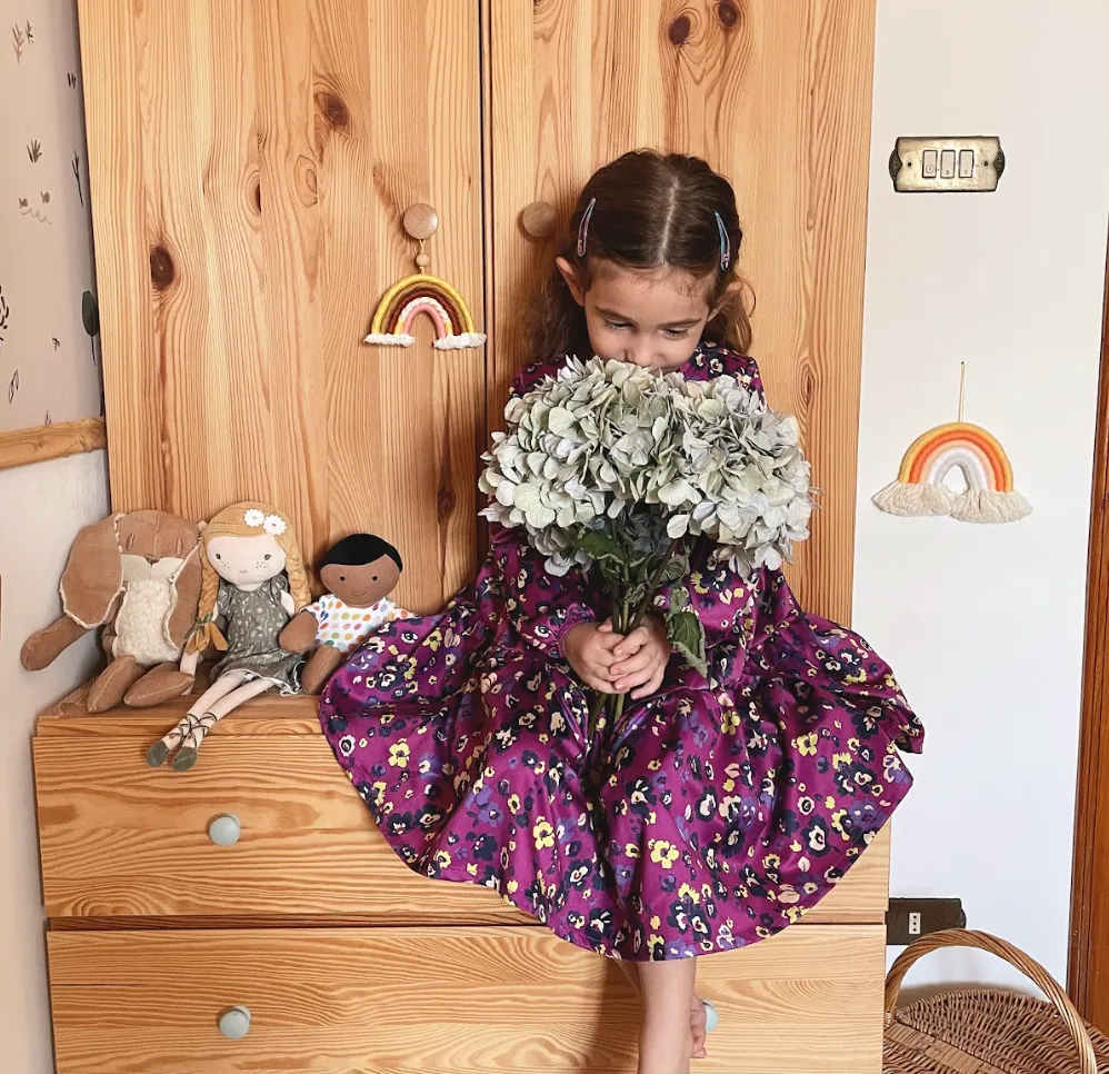 Una bambina con un vestito a fiori Melby tiene un mazzo di fiori, seduta su una cassettiera di legno